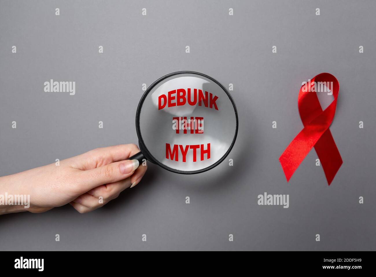 La main d'une femme tient une loupe avec du texte. Ruban rouge sur fond gris. Pose à plat. Le concept de la Journée mondiale du sida. Banque D'Images