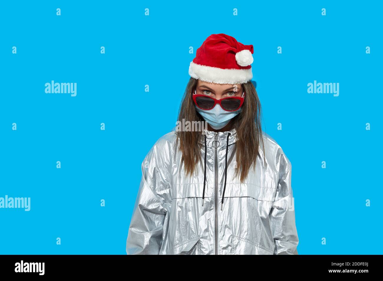Femme sérieuse portant chapeau de père Noël et masque de protection et lunettes de soleil rouges debout sur fond bleu en studio et regardant l'appareil photo. Lady regarde sur t Banque D'Images Femme sérieuse portant chapeau de père Noël et masque de protection et lunettes de soleil rouges debout sur fond bleu en studio et regardant l'appareil photo. Lady regarde sur t Banque D'Images