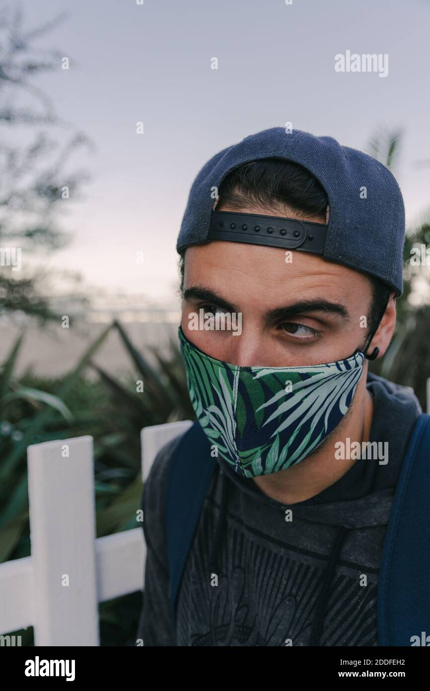 Photo verticale d'un jeune homme sud-américain portant un masque posé contre une clôture blanche Banque D'Images