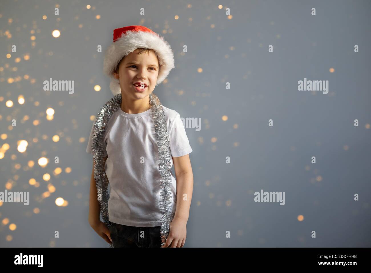 Joyeux garçon en T-shirt blanc et chapeau rouge de Noël sur fond gris avec lumières floues. Concept de Noël ou de nouvel an. Banque D'Images