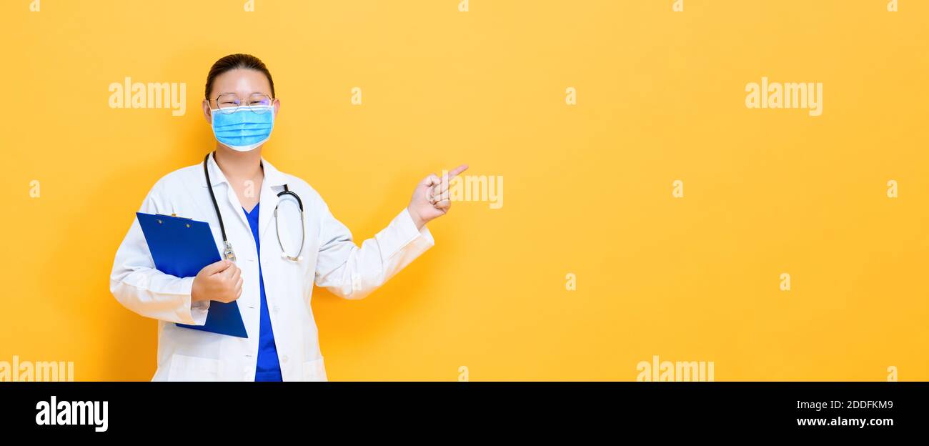 Jeune femme asiatique médecin portant un masque médical pointant de la main espace vide de côté isolé sur fond de bannière jaune Banque D'Images