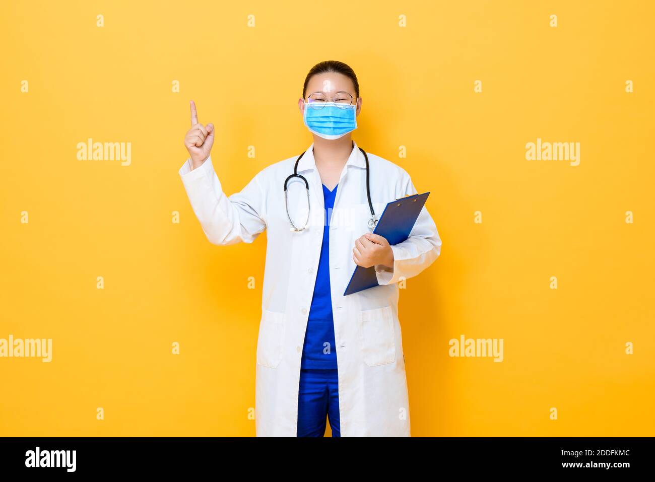 Jeune femme asiatique médecin portant un masque médical pointant de la main vers le haut pour vider l'espace isolé sur fond jaune Banque D'Images