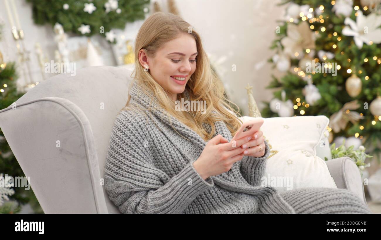 Femme souriante tendance dans un chandail gris confortable sittin sur confortable fauteuil et envoi de sms Banque D'Images
