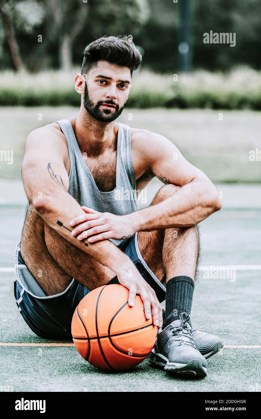 Tout le corps d'un jeune joueur de basket-ball à barbe musclé sportswear assis près du ballon sur le terrain de jeu de rue et regardant appareil photo Banque D'Images