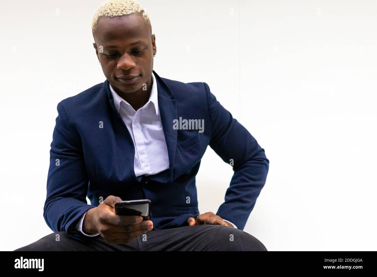 Homme d'affaires afro-américain souriant assis près d'un mur blanc et de messages sur un smartphone Banque D'Images