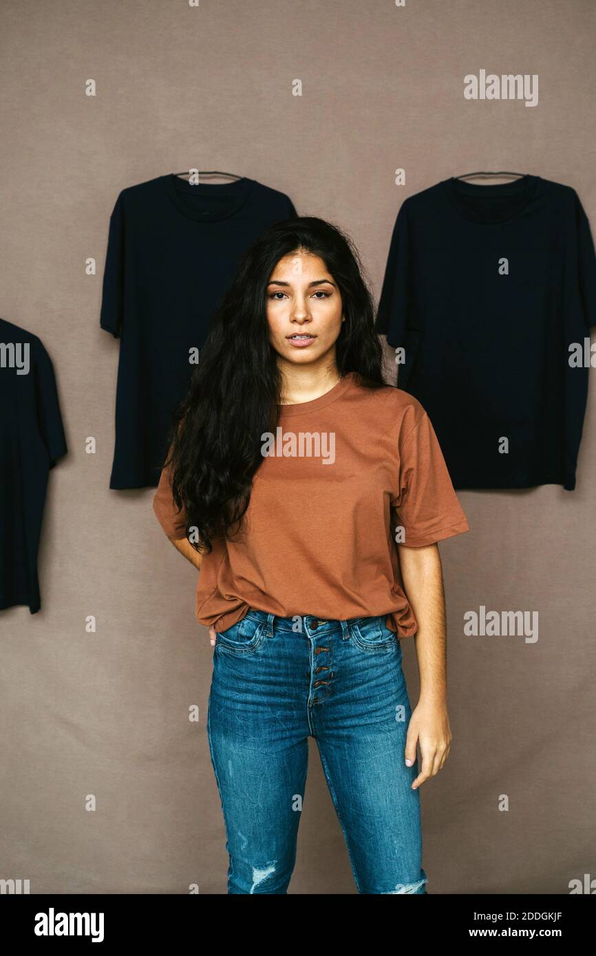 Femme ethnique dans une tenue décontractée debout en studio avec t des chemises sur le mur et regardant l'appareil photo Banque D'Images Femme ethnique dans une tenue décontractée debout en studio avec t des chemises sur le mur et regardant l'appareil photo Banque D'Images
