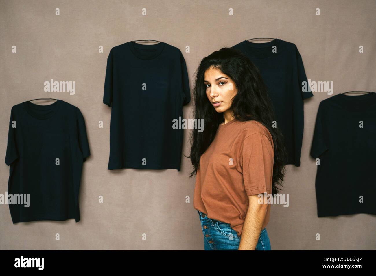 Vue latérale d'une femme ethnique dans une tenue décontractée studio avec t-shirts sur le mur et regardant la caméra Banque D'Images Vue latérale d'une femme ethnique dans une tenue décontractée studio avec t-shirts sur le mur et regardant la caméra Banque D'Images