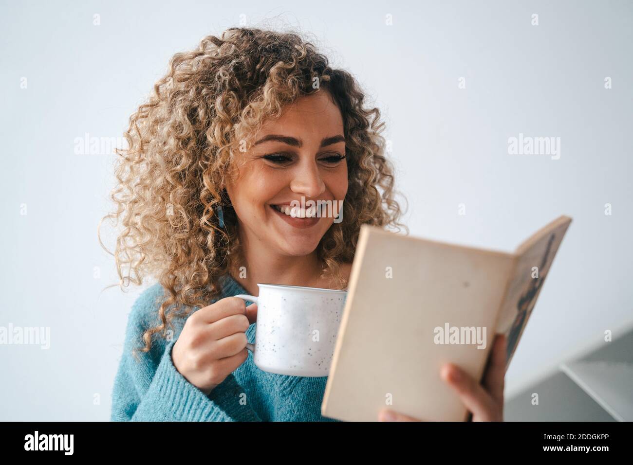 Une femme joyeuse dans un confortable chandail domestique penchée sur les escaliers à maison tout en appréciant l'histoire intéressante et en buvant un café chaud à week-end Banque D'Images