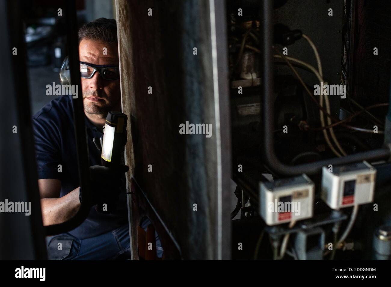 Mécanicien professionnel de lunettes de protection et d'écouteurs utilisant une lampe de poche lors de la vérification des détails de la machine pendant les travaux de réparation en atelier Banque D'Images