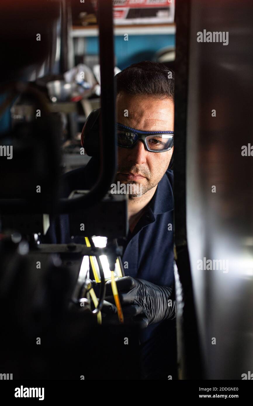 Mécanicien professionnel de lunettes de protection et d'écouteurs utilisant une lampe de poche lors de la vérification des détails de la machine pendant les travaux de réparation en atelier Banque D'Images