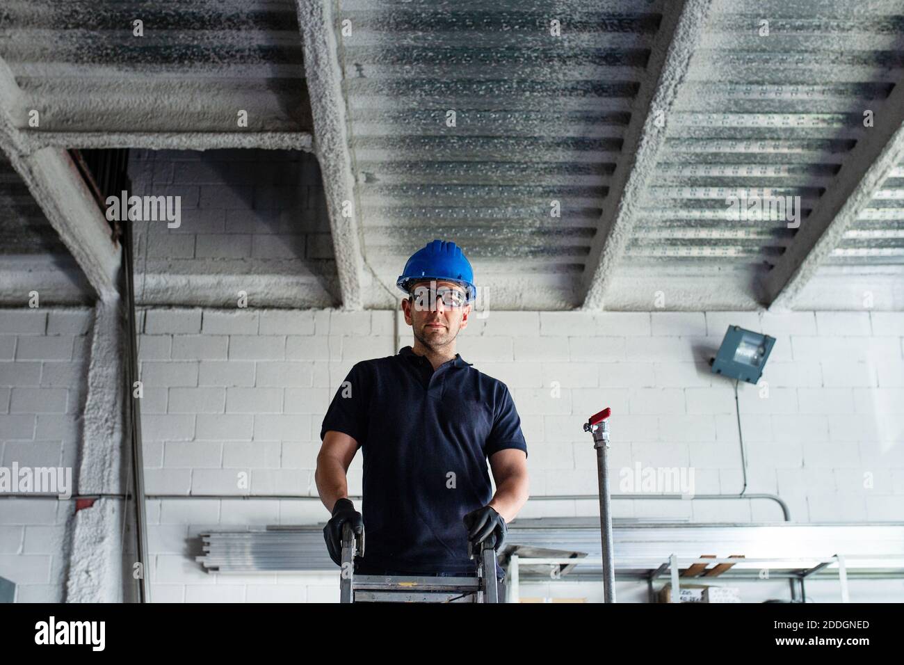 Faible angle d'homme joyeux ouvrier en casque de sécurité et de protection des lunettes se tenant sur l'échelle et en installant un tuyau métallique pendant le travail en atelier Banque D'Images