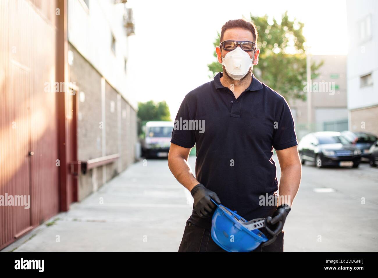 Adulte homme mécanicien en lunettes de protection et respirateur de maintien bleu hardhat entre les mains et regarde la caméra tout en se tenant à proximité atelier sur Banque D'Images