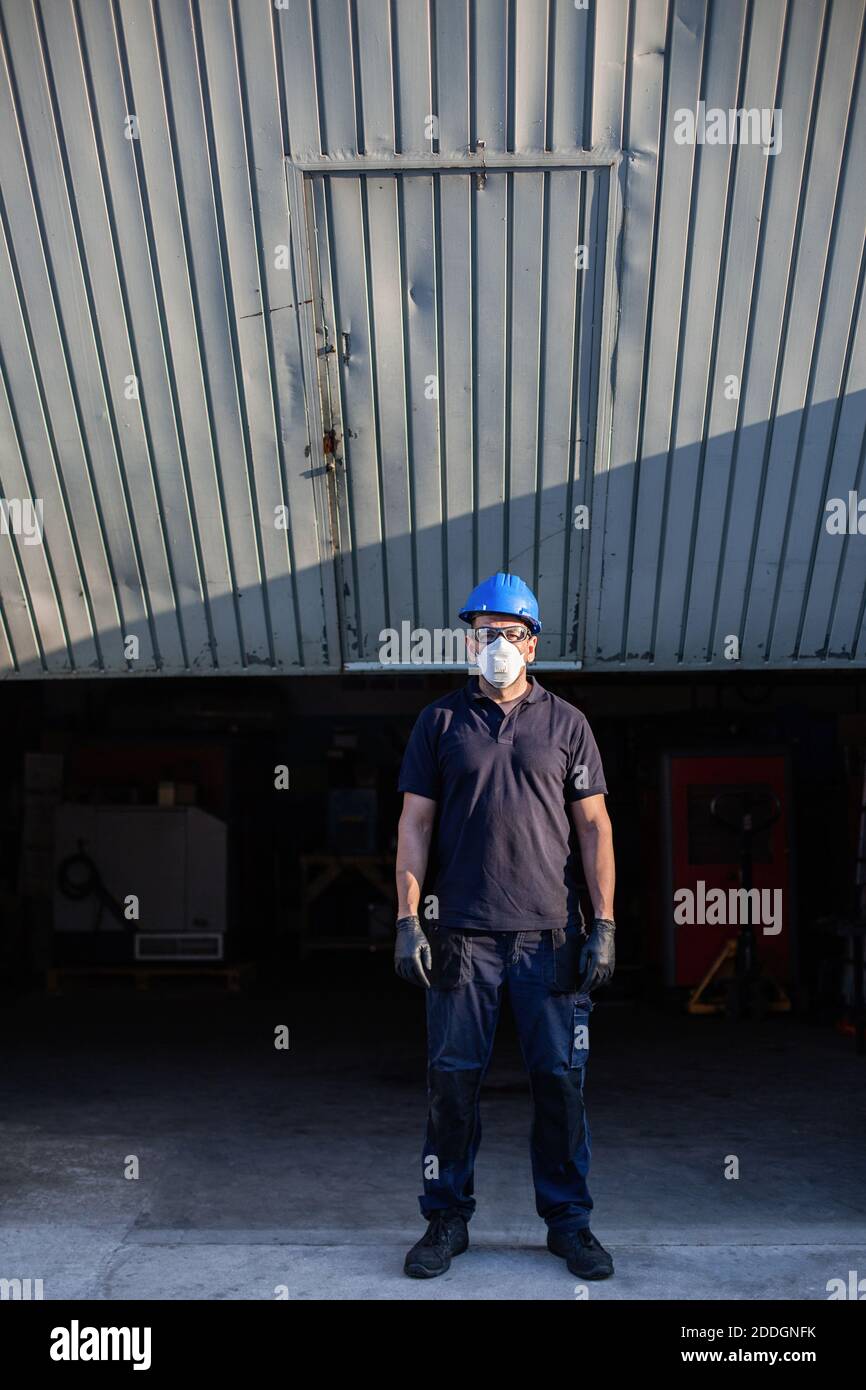 Adulte homme mécanicien en lunettes de protection et respirateur bleu hardhat regarde la caméra tout en se tenant près de l'atelier rue Banque D'Images