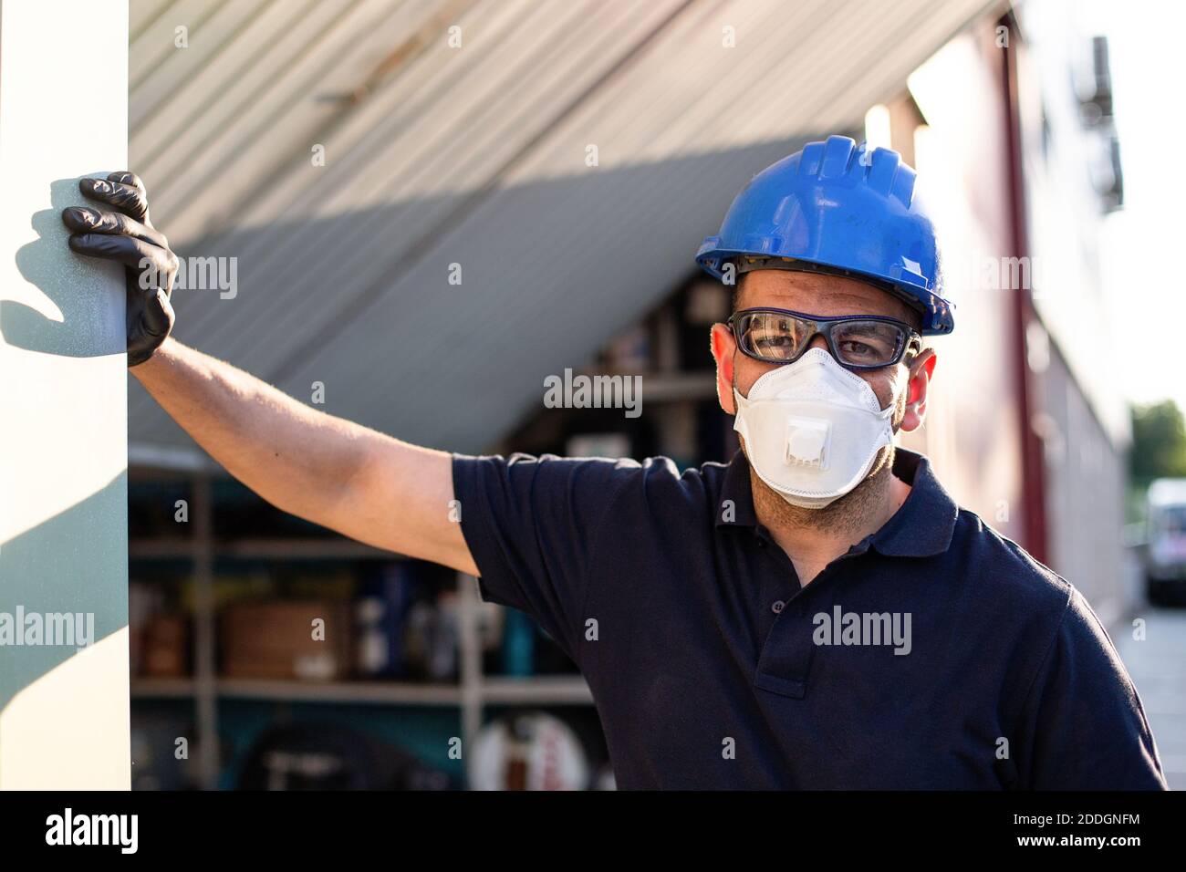 Adulte homme mécanicien en lunettes de protection et respirateur bleu hardhat regarde la caméra tout en se tenant près de l'atelier rue Banque D'Images