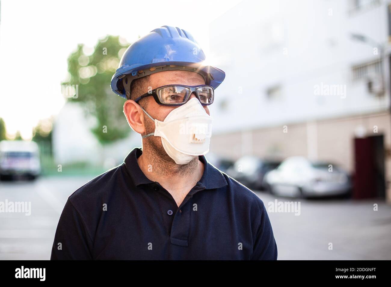 Adulte homme mécanicien en lunettes de protection et respirateur bleu hardhat se tient près du bâtiment de l'atelier dans la rue Banque D'Images