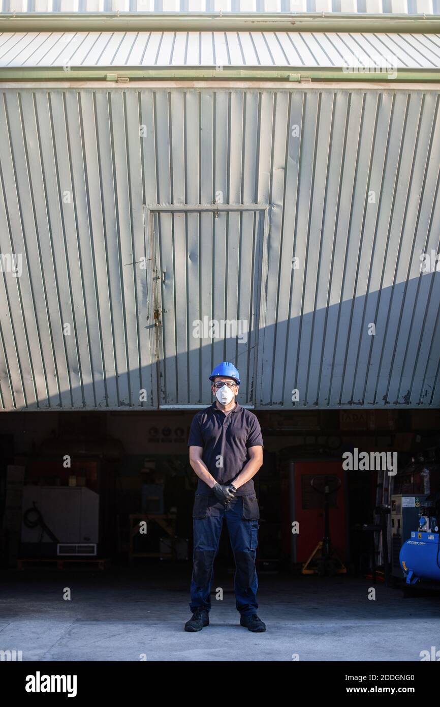 Adulte homme mécanicien en lunettes de protection et respirateur bleu hardhat regarde la caméra tout en se tenant près de l'atelier rue Banque D'Images