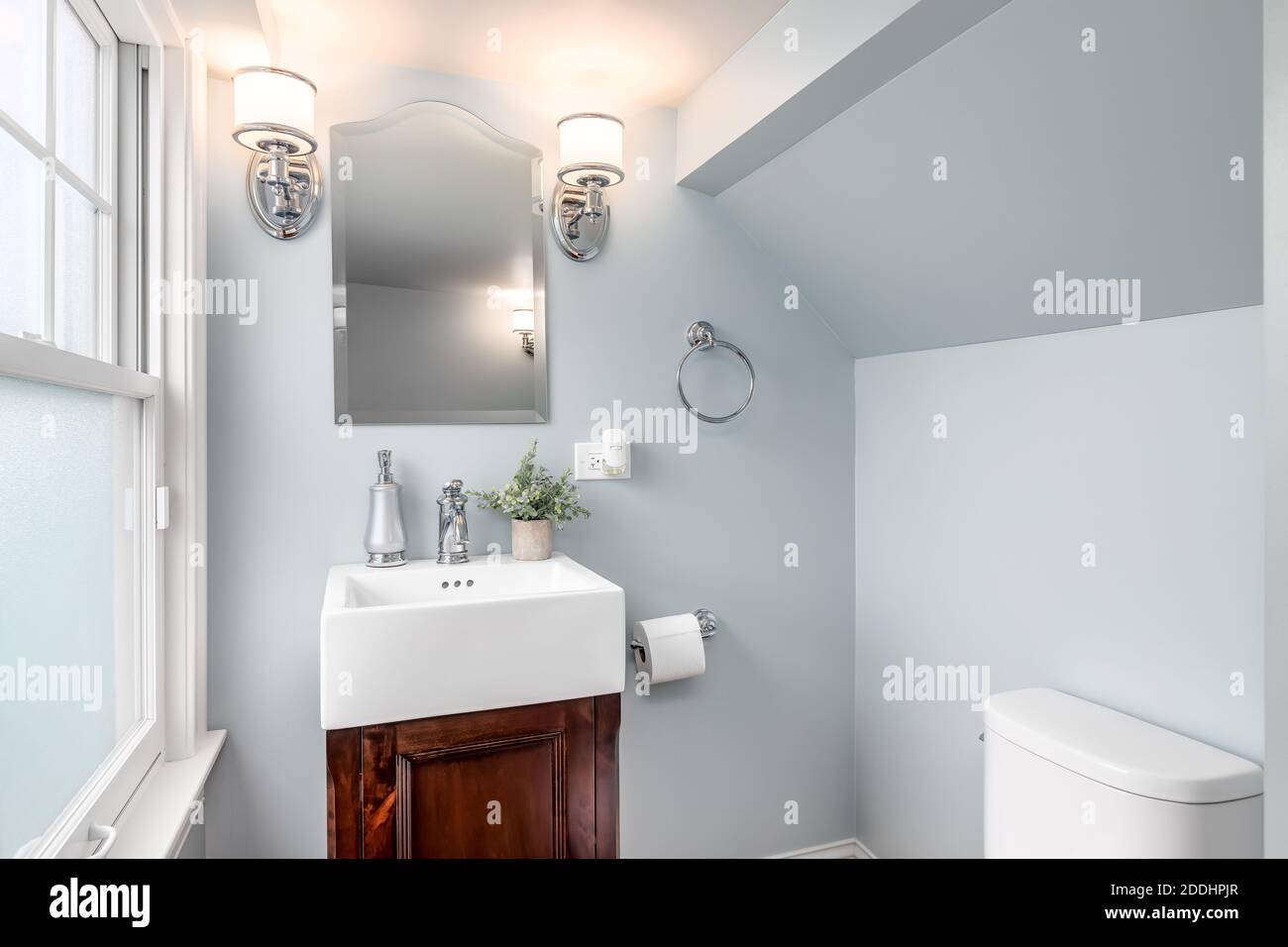 Une petite salle de bains avec des murs bleus, un meuble-lavabo en bois, des fleurs sur le lavabo blanc et des lumières montées à côté du miroir. Banque D'Images