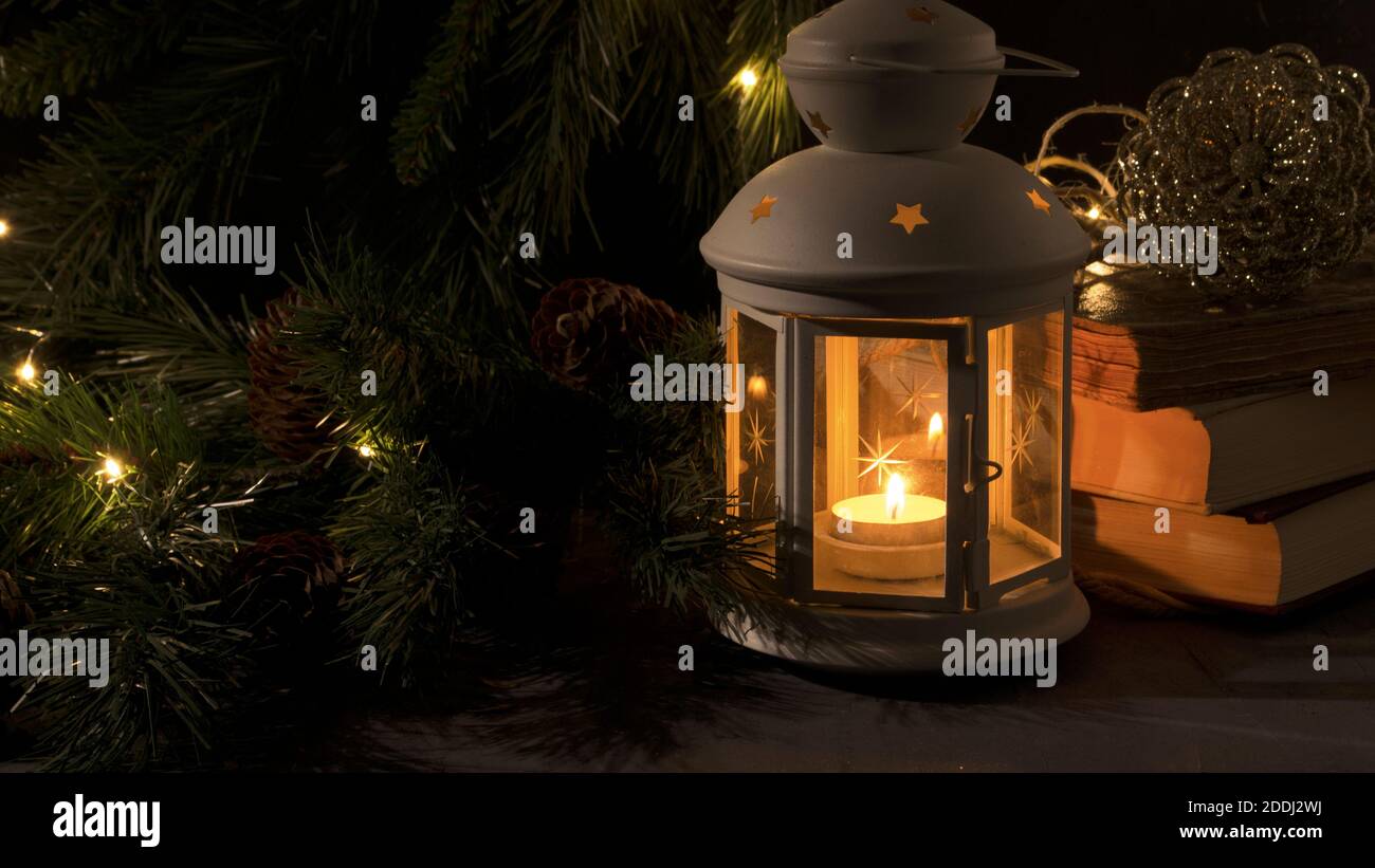Lanterne blanche avec une bougie allumée. Livres, branche d'arbre de Noël et lumières sur fond. Image sombre de nuit. Humeur mystère de Noël. Banque D'Images