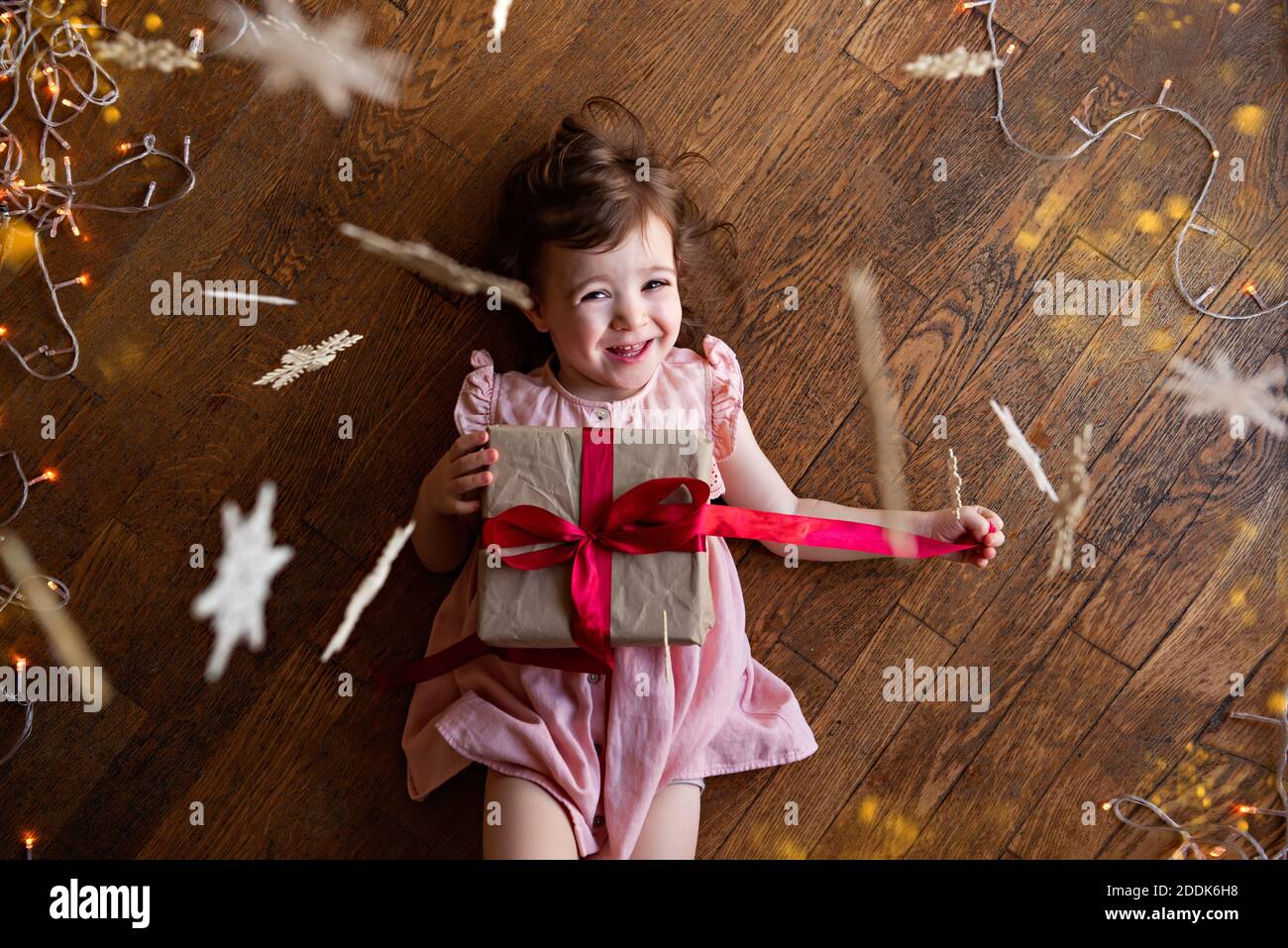 Petite fille heureuse dans une robe rose se trouve sur le plancher en bois. Contient une boîte de Noël dans un emballage artisanal, un cadeau de Noël avec un ruban rouge. Flocons de neige h Banque D'Images