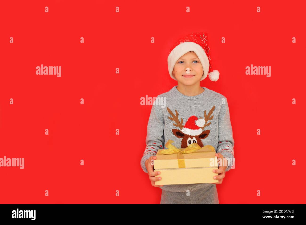 Un garçon dans un chapeau de père Noël et un chandail du nouvel an avec un cerf tiennent un cadeau du nouvel an sur fond rouge. L'enfant ressent les émotions de recevoir un cadeau de Noël Banque D'Images