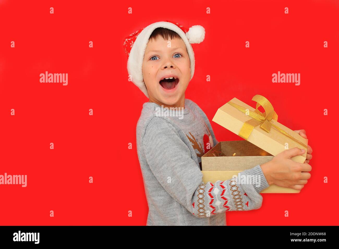 Un garçon dans un chapeau de père Noël et un chandail du nouvel an avec un cerf tiennent un cadeau du nouvel an sur fond rouge. L'enfant ressent les émotions de recevoir un cadeau de Noël Banque D'Images