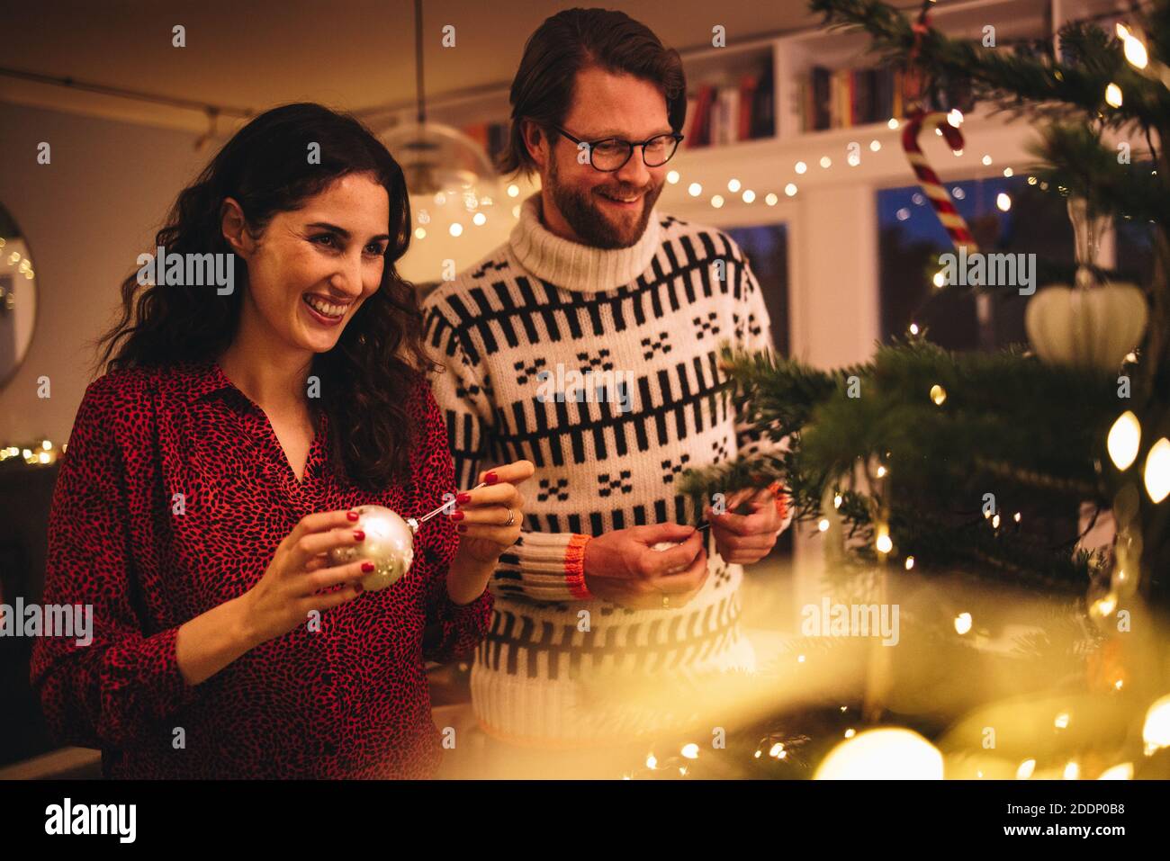 Femme nouant une boule décorative brillante sur l'arbre de Noël avec un homme debout près. Couple décorant arbre de Noël à la maison de famille. Banque D'Images
