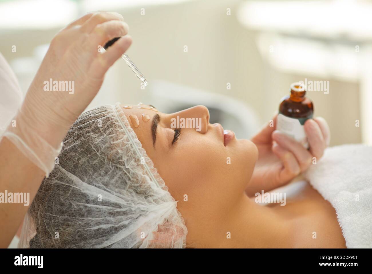 Mains de l'esthéticienne goutte d'huile nourrissante sur le front et le visage peau de la jeune femme Banque D'Images