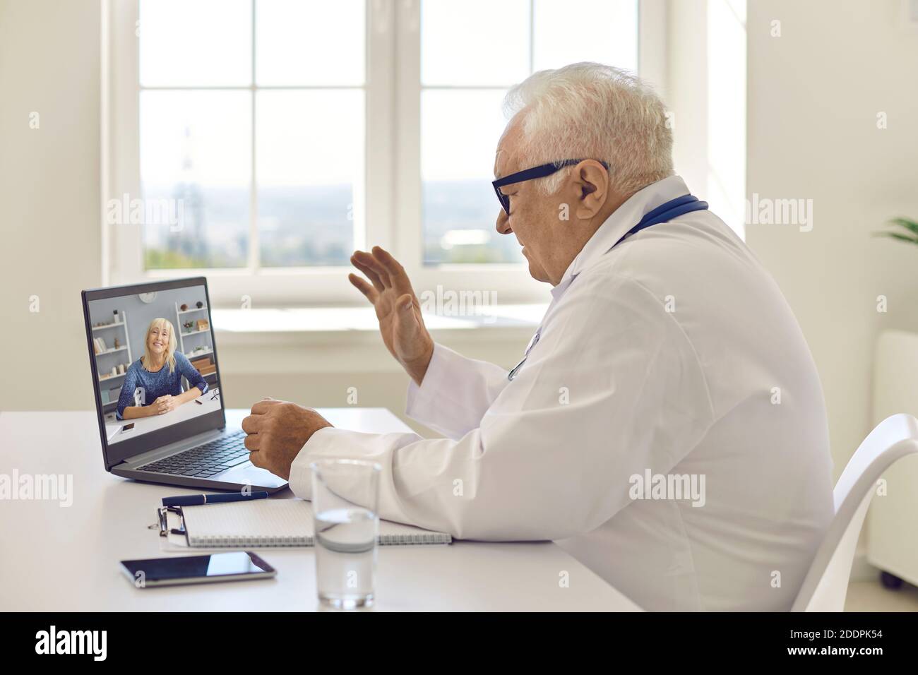 Médecin senior assis à un bureau, en agitant la main sur un ordinateur portable et en saluant le patient Banque D'Images