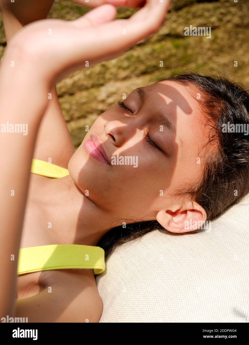 Jolie petite fille d'origine asiatique couchée sur un banc, prenant un bain de soleil le matin à la maison. Elle se lève la main pour couvrir son visage de la lumière intense du soleil Banque D'Images