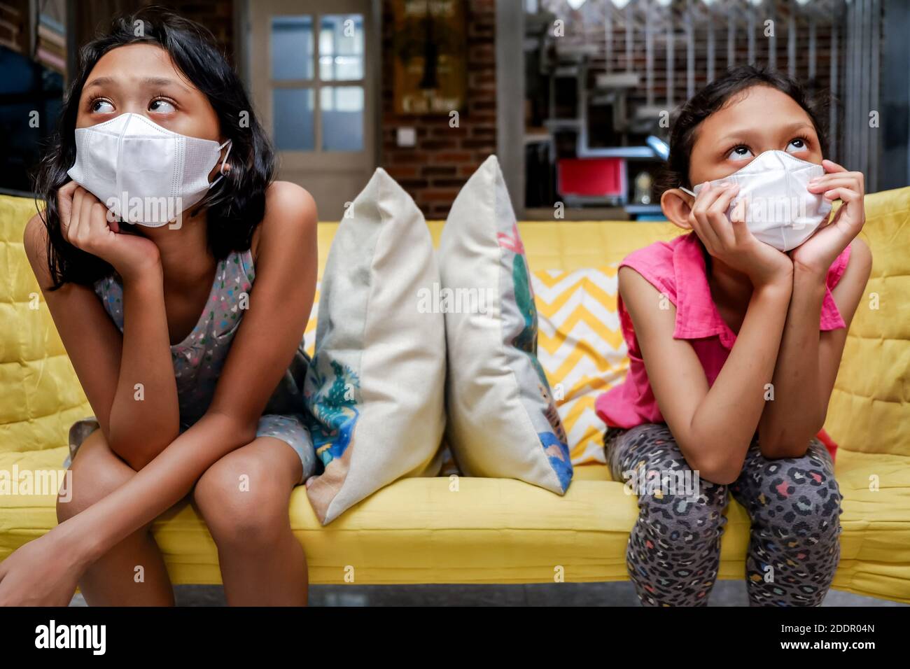 Deux petites filles d'Asie du Sud-est portant un masque médical pour le visage obtenir S'ennuient assis sur un canapé à part l'un de l'autre à la maison Pendant une pandémie Banque D'Images