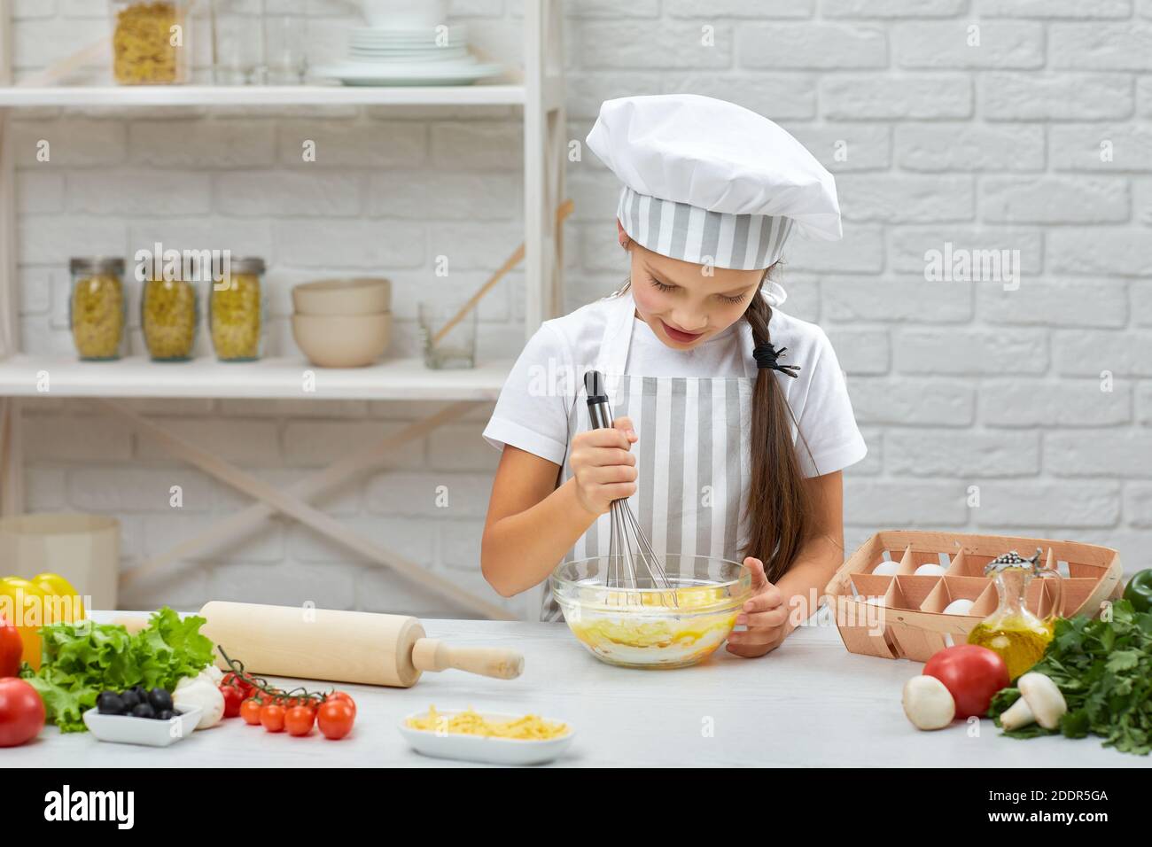 petite fille en chapeau et un tablier pétrissez avec l'acier fouetter la pâte dans la cuisine. Fouetter. Banque D'Images