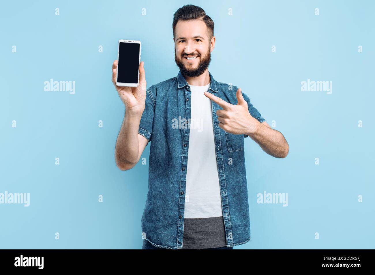 Jeune homme heureux tenant un smartphone dans sa main et montrant un écran de smartphone vierge à l'appareil photo, pointant vers lui avec son doigt, sur un bac bleu Banque D'Images