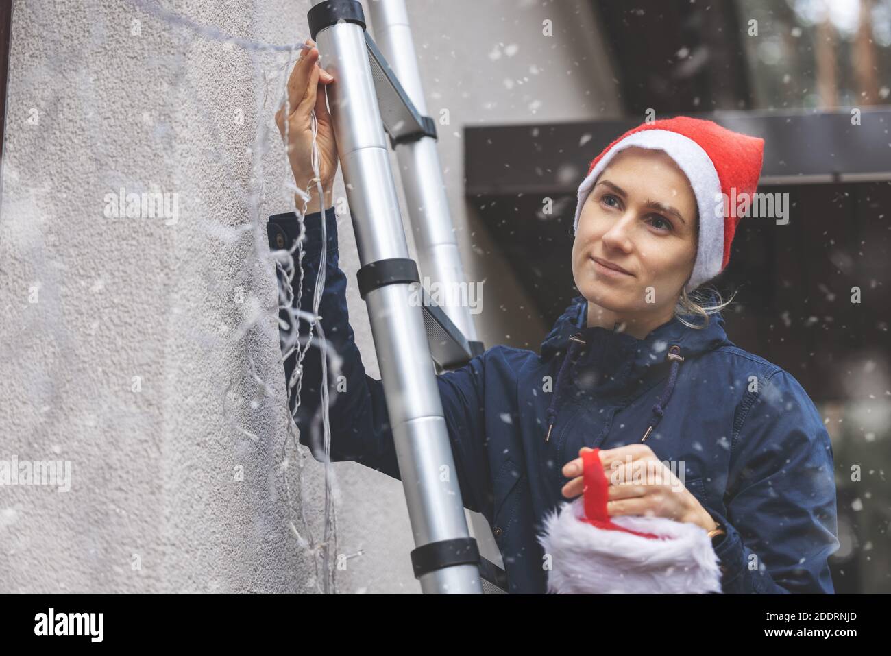 femme avec chapeau de père noël debout sur l'échelle à l'extérieur et l'installation lumières et décors de noël sur la façade de la maison Banque D'Images