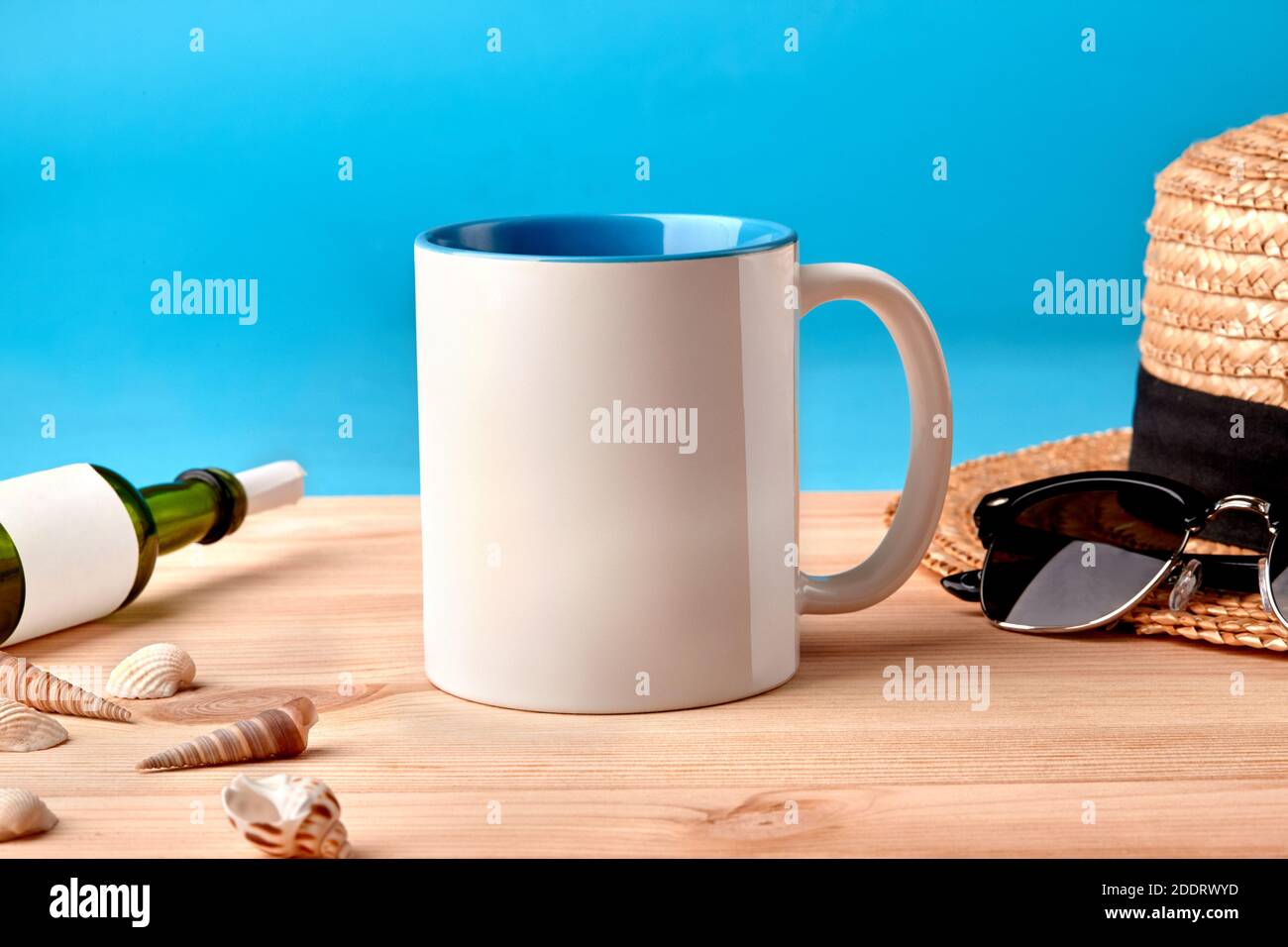Mug en céramique blanche sur un bureau en bois à côté des coquillages, bouteille avec feuille de papier enroulée, chapeau de paille et lunettes de soleil noires, fond bleu Banque D'Images