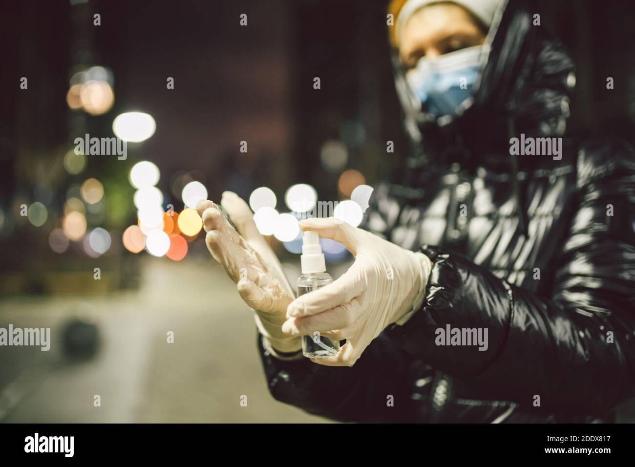 Femme mature avec masque facial et gants médicaux en caoutchouc tient antiseptique tout en se tenant contre la ville de nuit par temps froid. La femme âgée utilise la main Banque D'Images