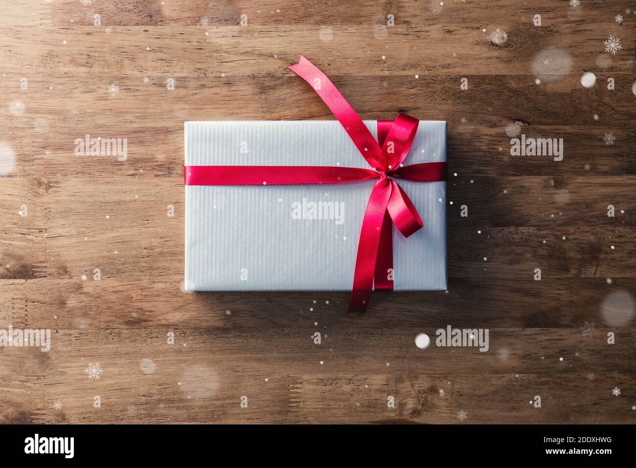 Boîte cadeau de Noël et du nouvel an emballée d'argent papier gris et ruban rouge noeud sur table en bois - vue de dessus avec effet de chute de neige Banque D'Images