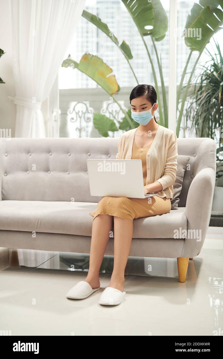 Belle jeune femme asiatique assise sur un canapé portant un masque médical, travail à la maison avec ordinateur portable pendant le coronavirus Banque D'Images