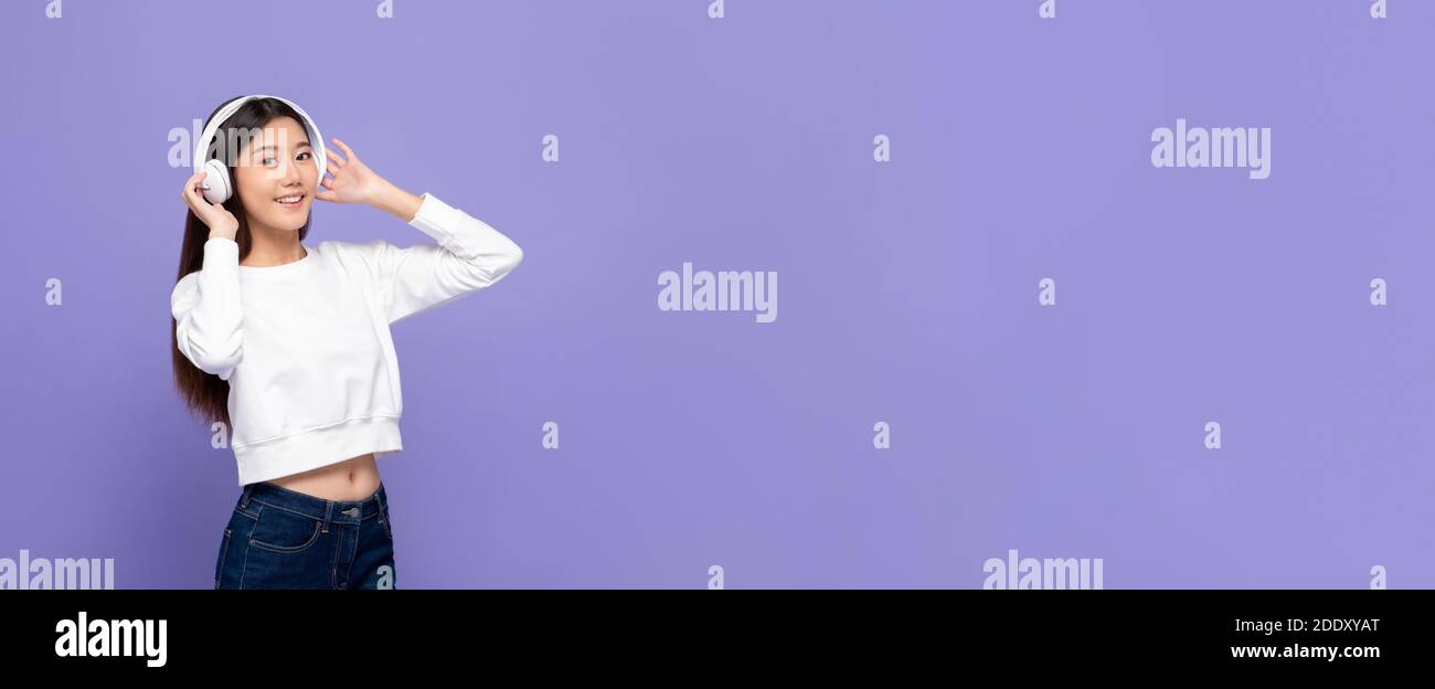 Jeune femme asiatique mignonne en souriant et en touchant des écouteurs écouter de la musique à côté de l'espace de copie isolé sur une bannière violette arrière-plan Banque D'Images