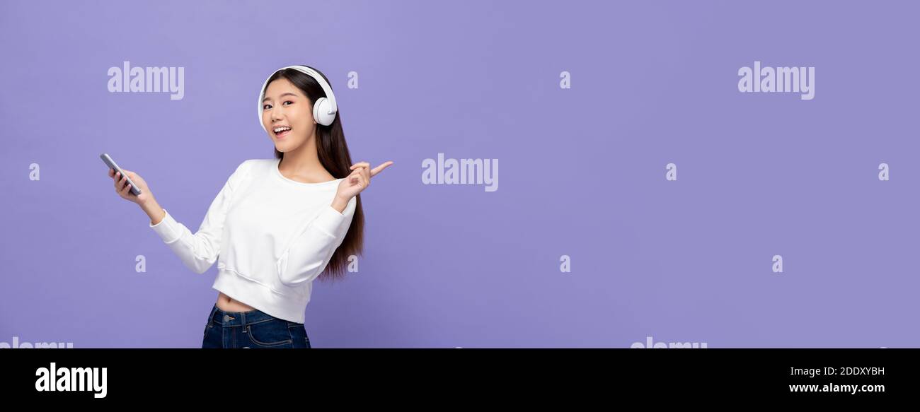 Jeune femme asiatique portant un casque qui écoute de la musique téléphone mobile et pointage vers l'espace de copie sur violet clair arrière-plan de la bannière Banque D'Images