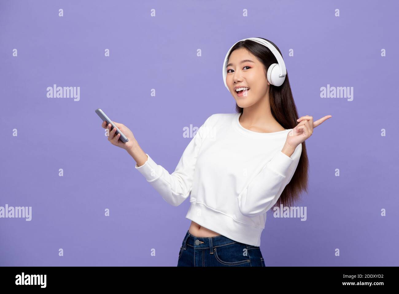 Jeune femme asiatique portant un casque qui écoute de la musique smartphone et pointage sur fond violet clair Banque D'Images