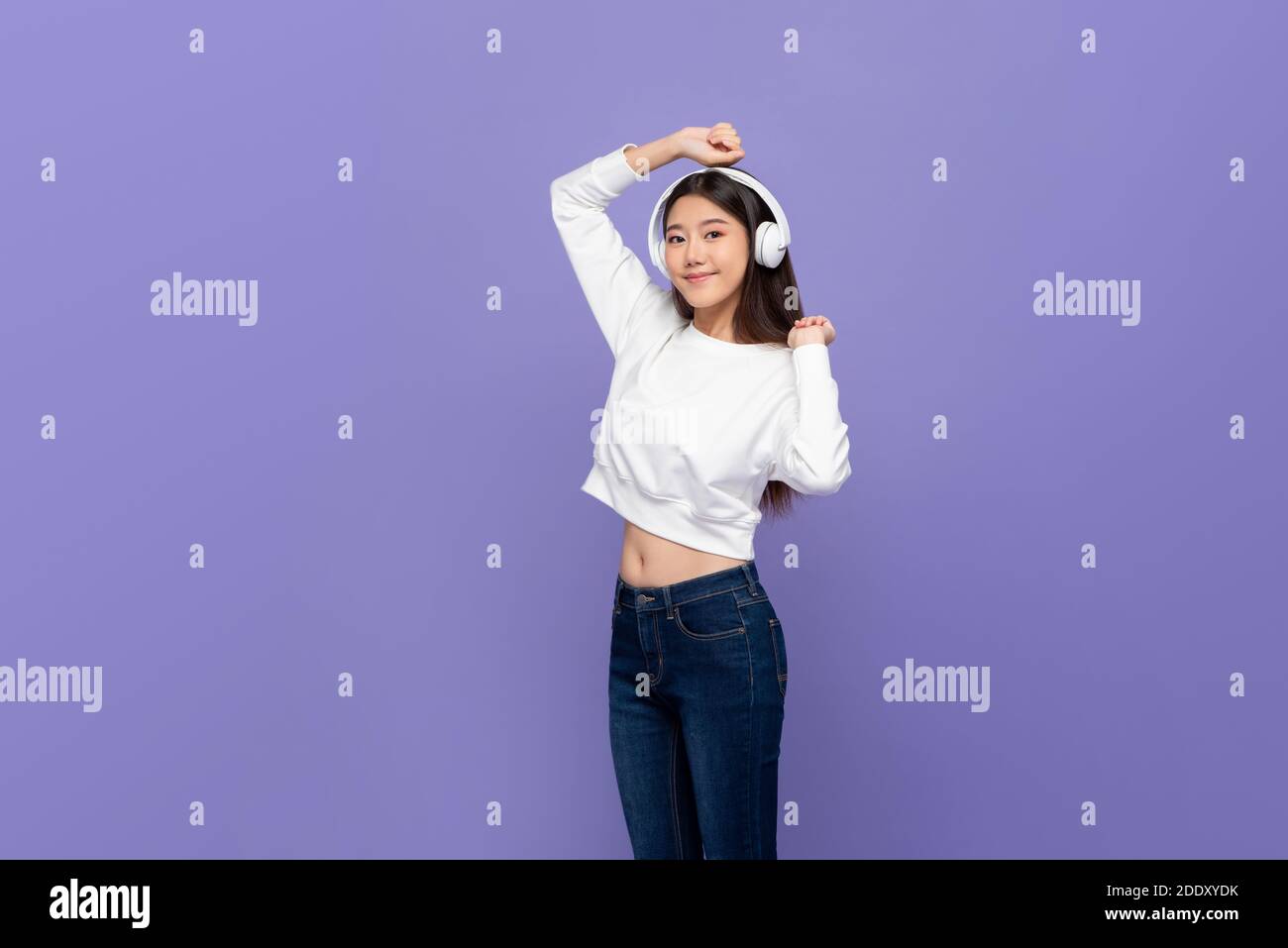 Jeune femme asiatique portant un casque sans fil pour écouter de la musique et danser sur fond violet clair Banque D'Images