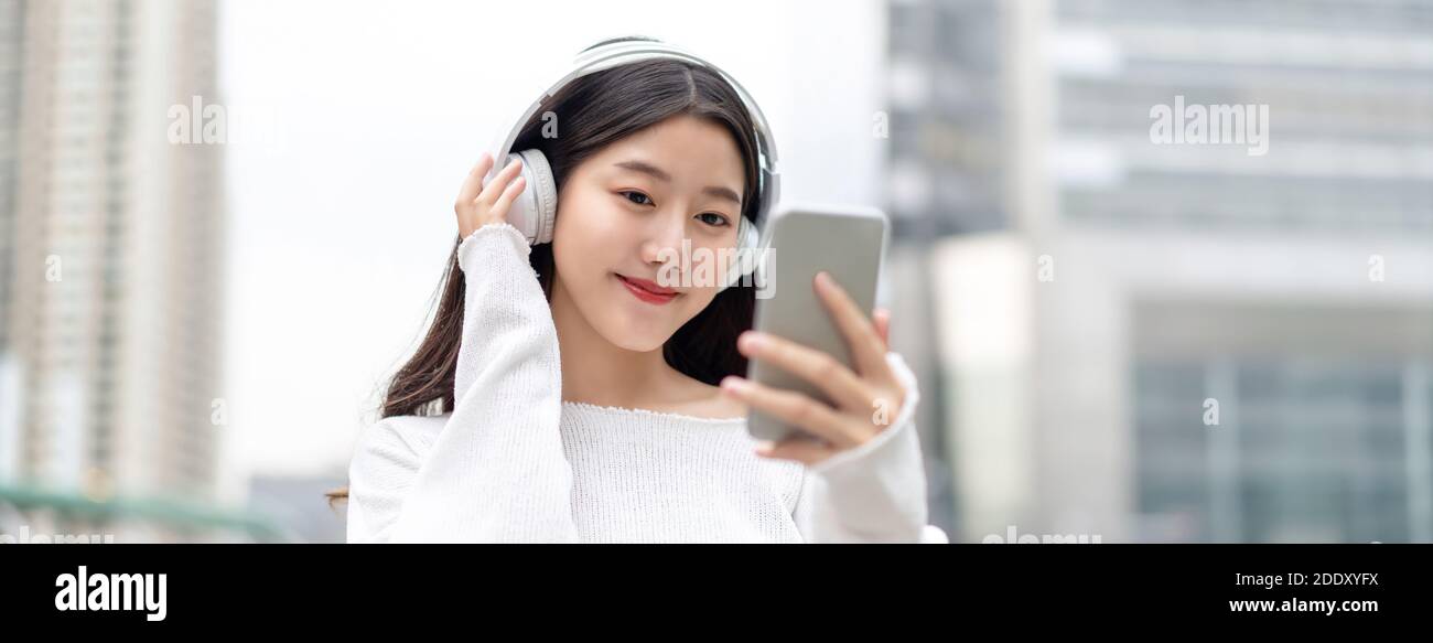 Bannière de jeune heureuse jolie femme asiatique mignon regarder et écouter de la musique en streaming tout en souriant et en touchant casque contre la cit Banque D'Images
