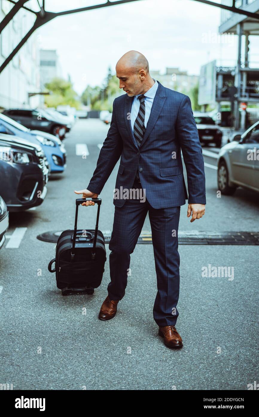 Homme d'affaires chauve marchant sur le parking de l'aéroport avec des valises et des voitures garées. Homme d'affaires en costume et valise lors d'un voyage d'affaires. Homme marchant Banque D'Images