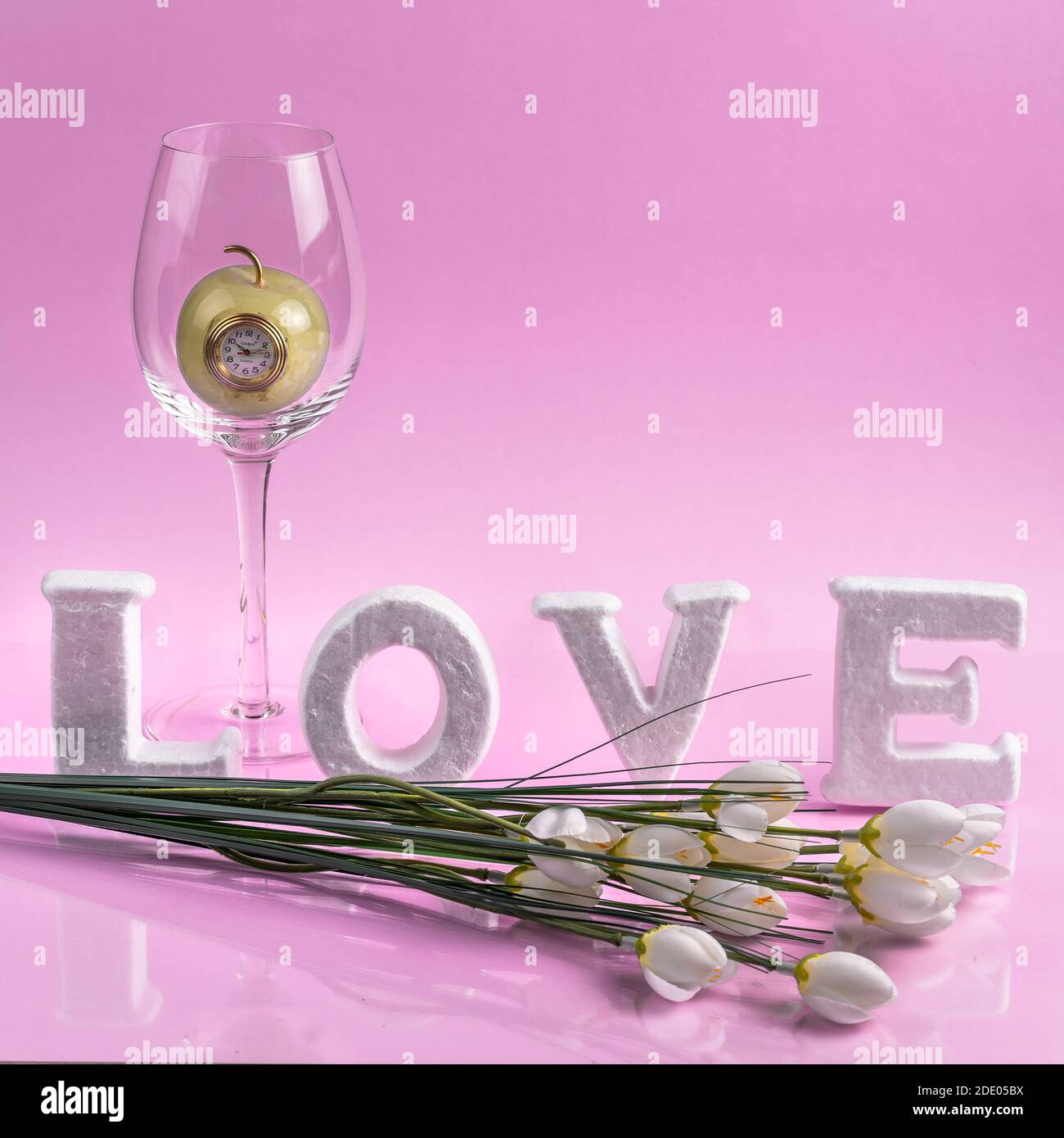 Idée de concept de la Saint-Valentin créative avec des lettres. Un verre avec une horloge de pomme à l'intérieur et des fleurs devant des lettres. Fond rose. Banque D'Images