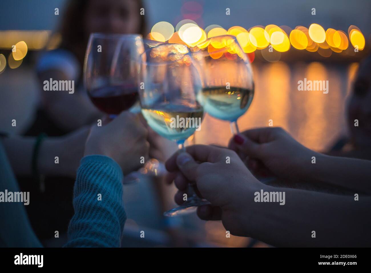 une fête à l'extérieur, célébrant et élevant des verres avec du vin contre le ciel bleu de nuit et les lumières de la ville jaune, le bokeh agréable et les réflexions de lumières dans l'eau Banque D'Images