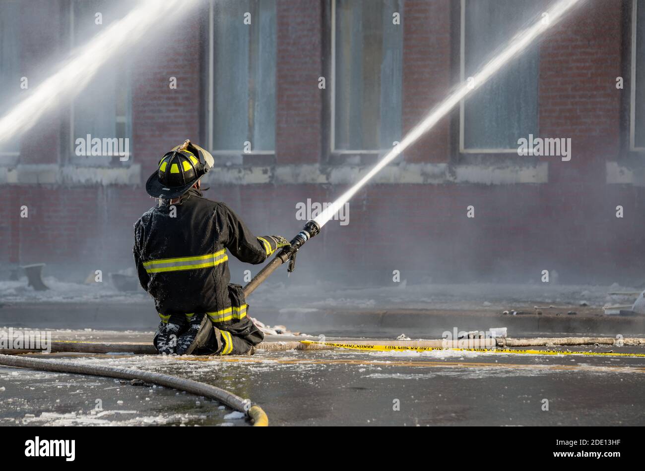 Un pompier s'agenouille au milieu d'une route mouillée et glacée. Il utilise un tuyau pour vaporiser de l'eau. Les tuyaux d'incendie sont au sol. Le bâtiment est en arrière-plan. Banque D'Images
