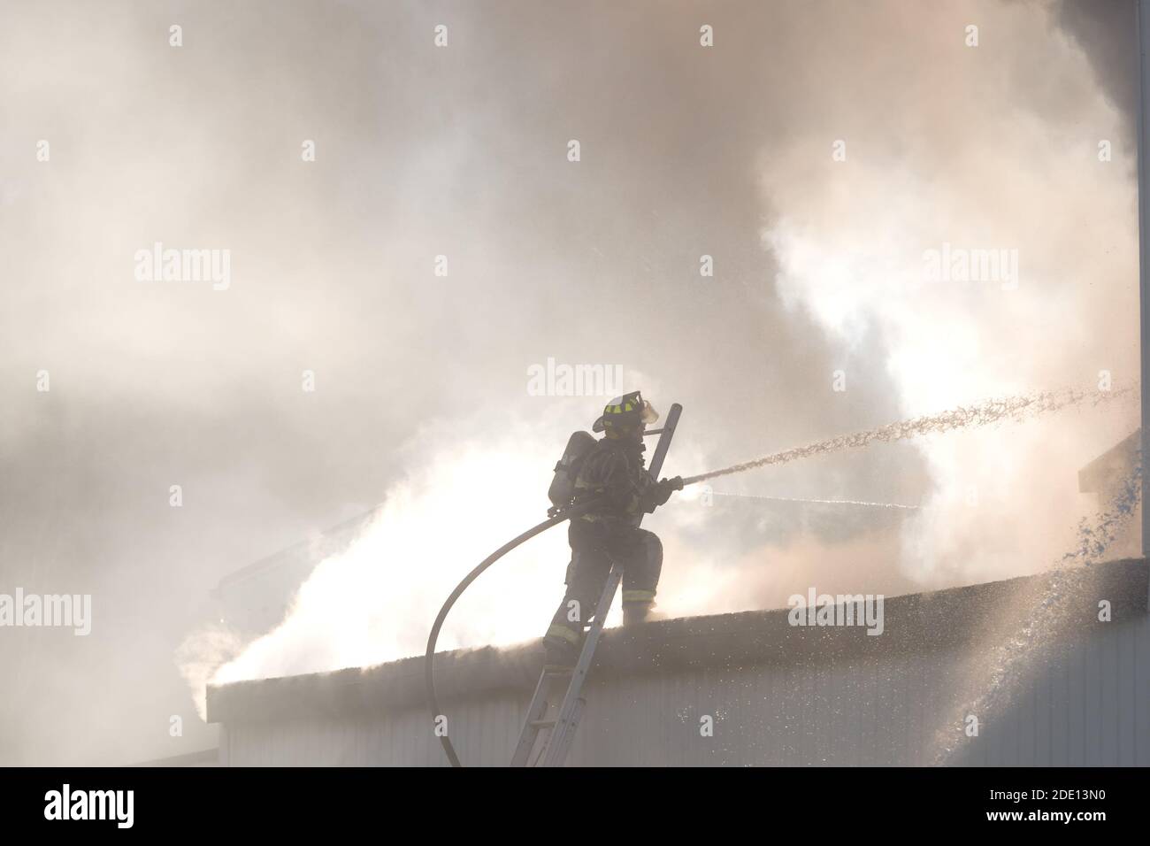 Pompier combattant un incendie avec un tuyau au sommet d'une échelle. Il est partiellement obscurci par la fumée. Beaucoup de pulvérisation et de fumée remplissent l'air. Espace pour le texte ci-dessus. Banque D'Images
