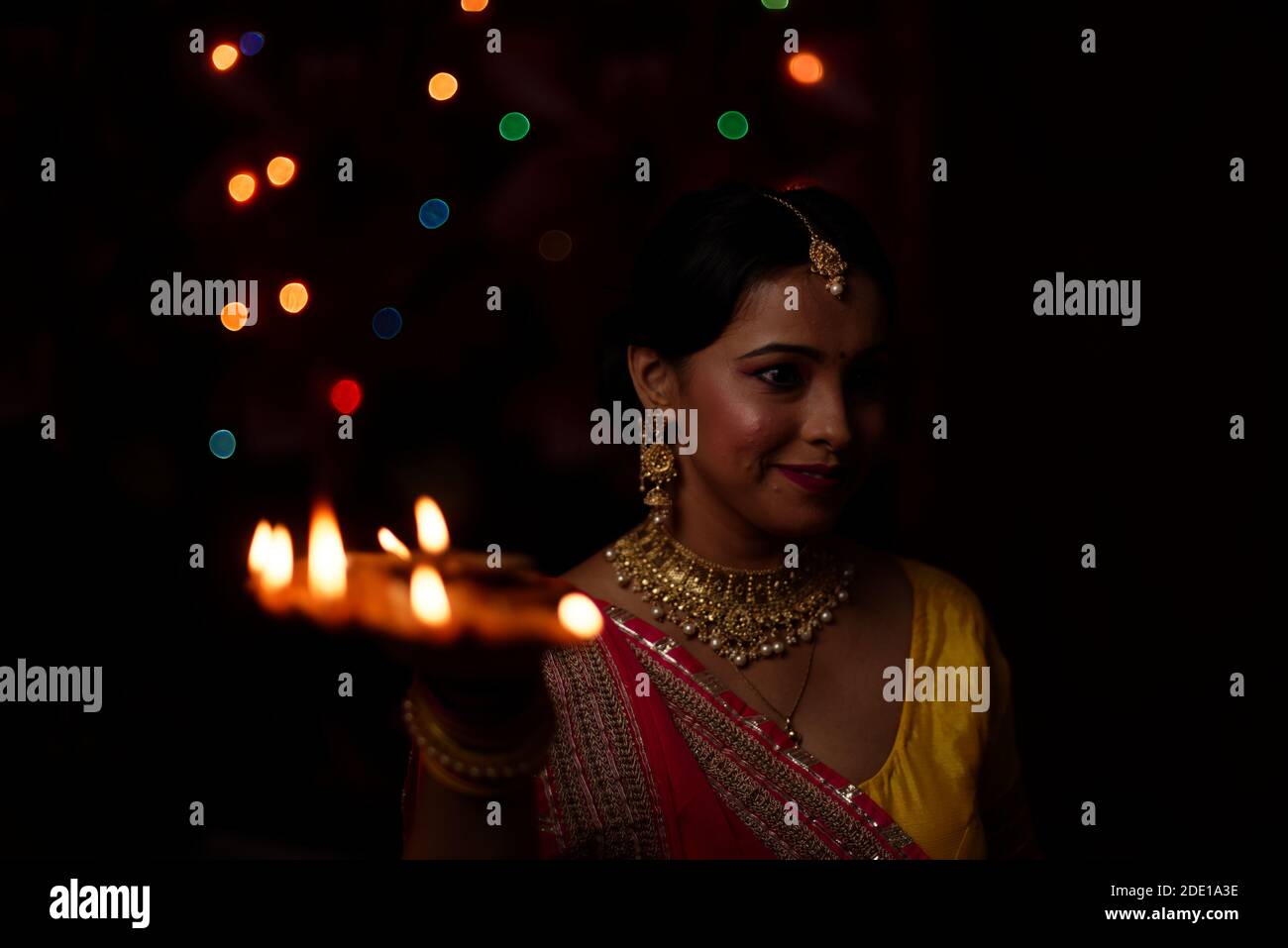 Une jeune et belle femme bengali indienne en robe traditionnelle indienne tient une diya/lampe Diwali dans sa main devant des lumières bokeh colorées. Banque D'Images