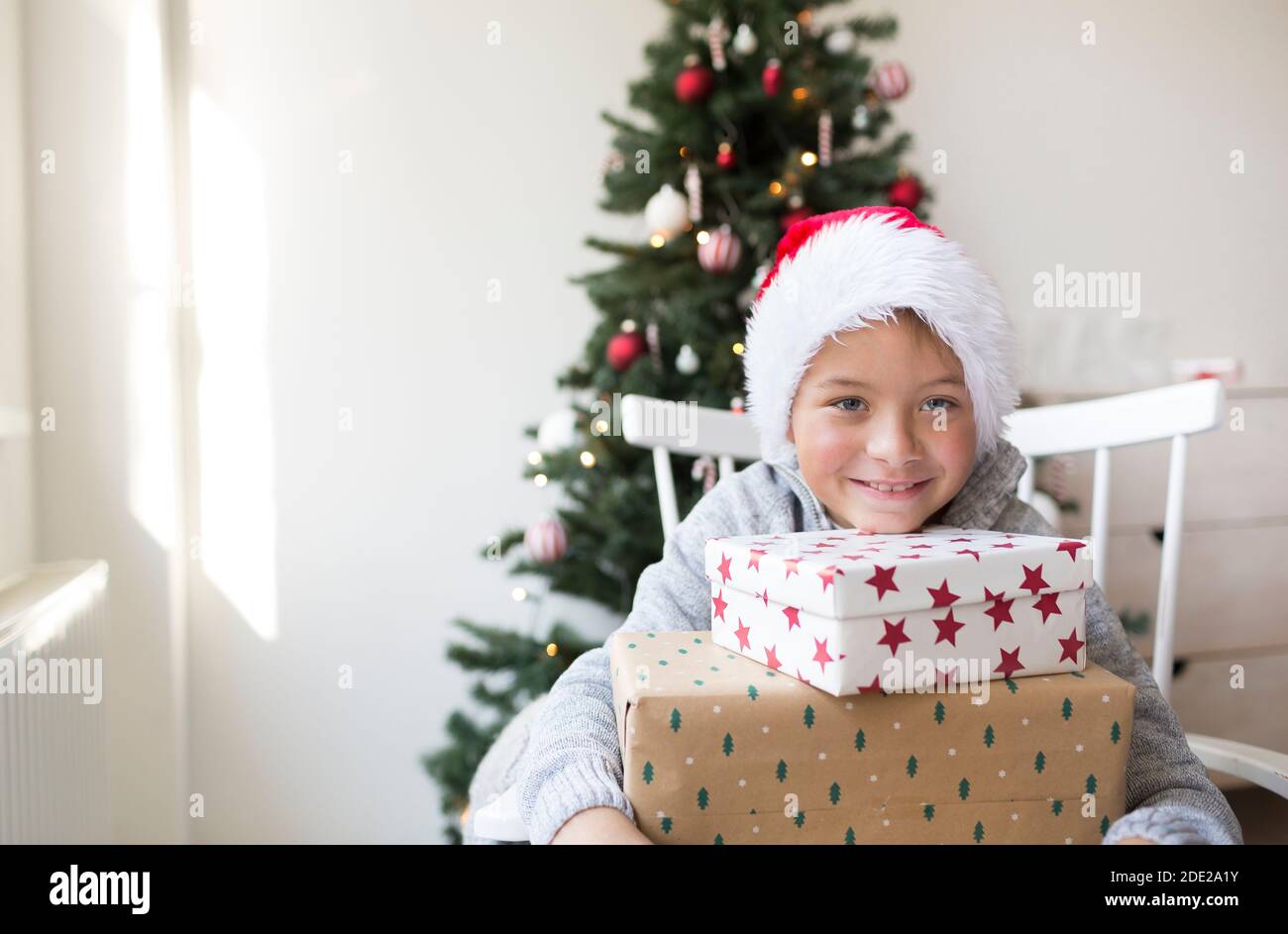 Jeune garçon dans la clause santa chapeau devant un Arbre de Noël avec cadeaux de Noël Banque D'Images