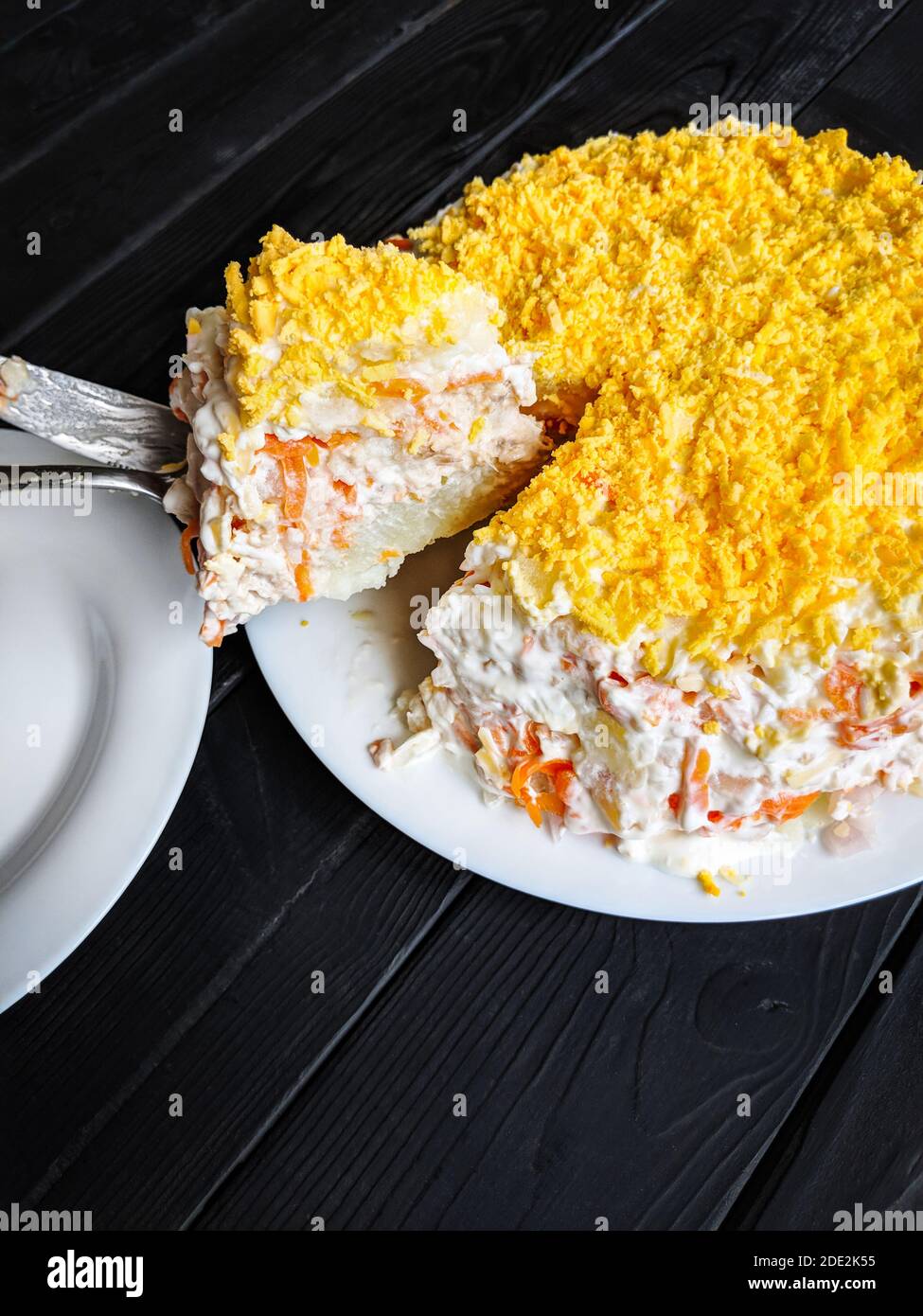 Salade russe maison Mimosa avec des pommes de terre, du poisson, des carottes, de la mayonnaise et râpez des jaunes d'œufs dans une assiette sur une table en bois sombre. Une tranche a été coupée et moi Banque D'Images